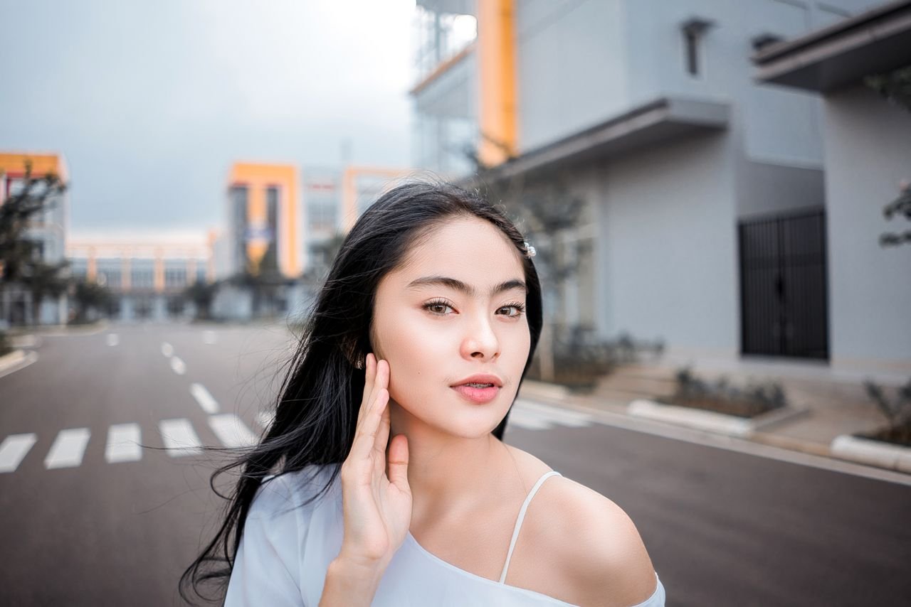 Self esteem young ethnic female with long black hair touching cheek and looking at camera while standing on asphalt road in modern city district